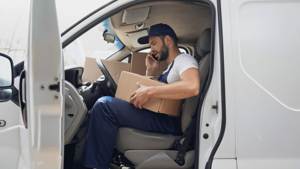Side view of courier talking on smartphone near cardboard boxes in car  - Zdjęcie, obraz