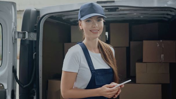 Cheerful delivery woman holding smartphone and looking at camera near blurred boxes in car outdoors  - Foto, Bild