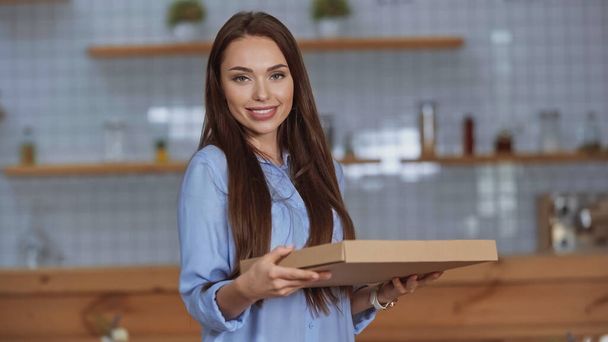 Mosolygó barna nő nézi a kamerát, miközben tartja pizza doboz otthon  - Fotó, kép