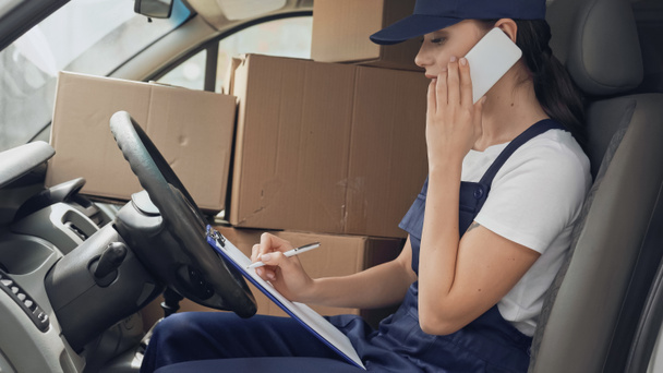 Vista lateral de la mujer de la entrega hablando en el teléfono inteligente y la escritura en el portapapeles cerca de cajas en el coche  - Foto, imagen