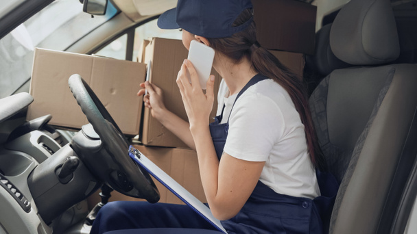 Delivery woman talking on cellphone near carton boxes and clipboard in car  - Zdjęcie, obraz