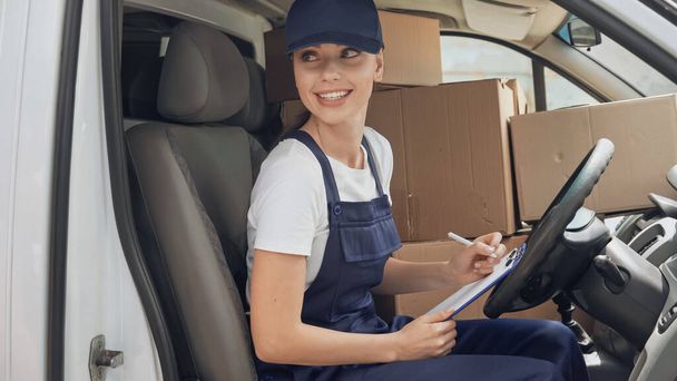 Cheerful delivery woman in uniform writing on clipboard near carton boxes in car  - Фото, зображення