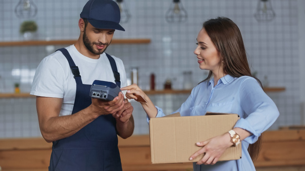Smiling woman holding carton box and using payment terminal near delivery man at home  - Фото, изображение
