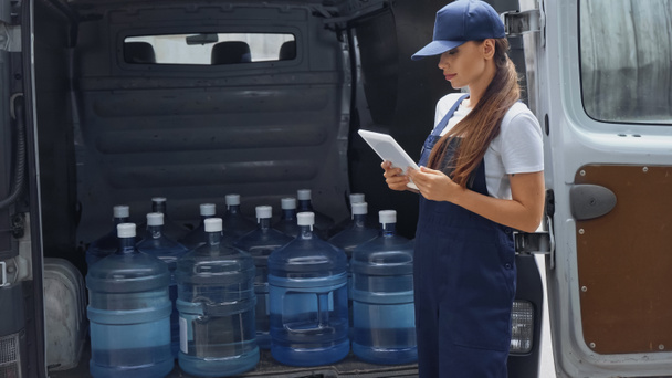Brunette delivery woman using digital tablet near bottles with water in car outdoors  - Фото, изображение