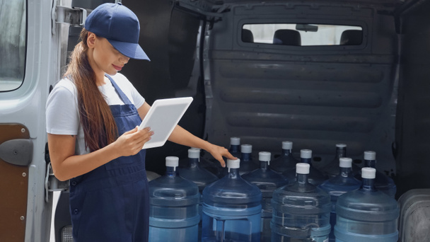 Delivery woman in overalls holding digital tablet near bottles with water in car outdoors - Foto, immagini