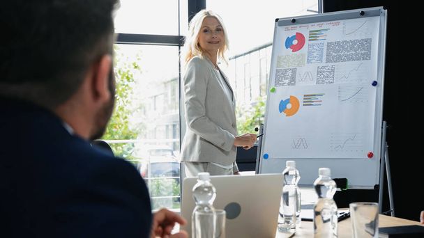 blonde middle aged businesswoman standing near flipchart with graphs and smiling to blurred colleague - Фото, изображение
