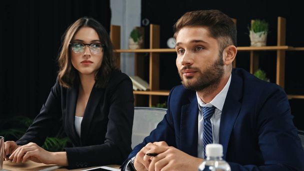 serious brunette businesswoman in eyeglasses looking at young colleague in office - Photo, Image