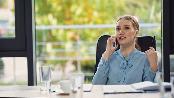 smiling businesswoman talking on cellphone near blurred notebook and beverages - Foto, Bild