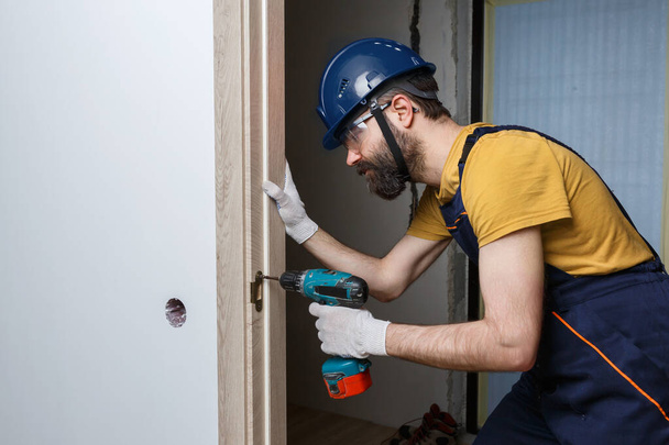 Ein Arbeiter mit einem Bohrer in einem orangefarbenen Helm installiert eine Tür im Haus - Foto, Bild