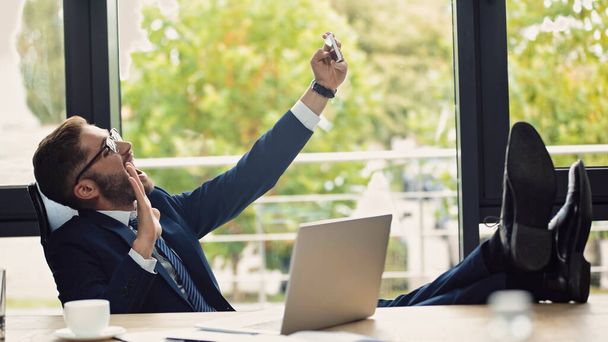ενθουσιασμένος επιχειρηματίας κάθεται με τα πόδια στο γραφείο και χαιρετώντας το χέρι, ενώ λαμβάνοντας selfie στο smartphone - Φωτογραφία, εικόνα