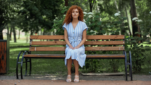 full length of redhead woman in blue striped dress sitting on bench in park - Foto, Imagen