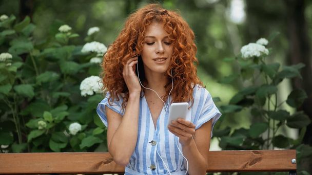 boldog fiatal vörös hajú nő hallgat zenét fülhallgató és okostelefon zöld parkban  - Fotó, kép