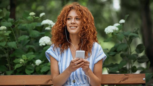 pleased redhead woman listening music in earphones and using smartphone in park  - Foto, Bild
