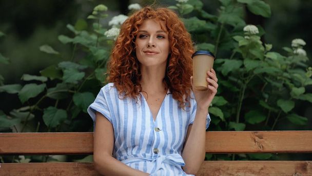smiling and redhead woman in dress holding paper cup while sitting on bench in park  - Photo, Image
