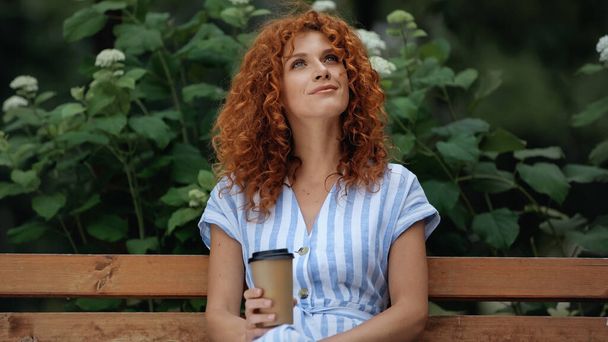 positive redhead woman in dress holding paper cup while sitting on bench in park  - Photo, image