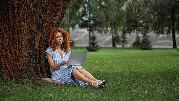 full length of curly redhead woman in dress listening music while using laptop in park  - Fotoğraf, Görsel