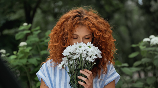 curly redhead woman with closed eyes smelling white flowers  - Фото, изображение