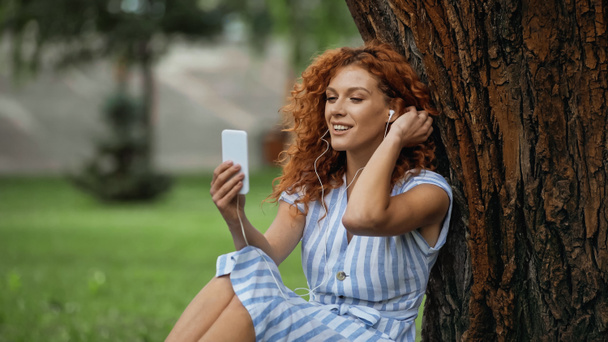 tevreden roodharige vrouw in blauwe jurk en oortelefoons luisteren muziek terwijl het kijken naar smartphone  - Foto, afbeelding