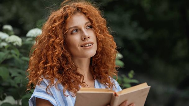 dreamy redhead woman smiling while holding book outdoors - Photo, Image