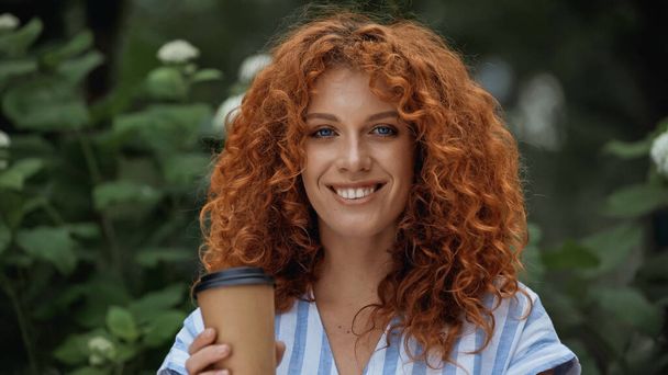 cheerful redhead woman holding paper cup outdoors - Foto, afbeelding