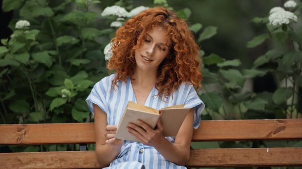 curly redhead woman smiling while reading book and sitting on wooden bench in park  - 写真・画像