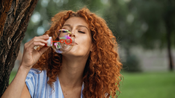 curly redhead woman blowing soap bubble in park  - Foto, imagen