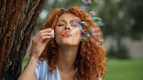 curly redhead woman frowning while blowing soap bubbles in park  - Photo, Image
