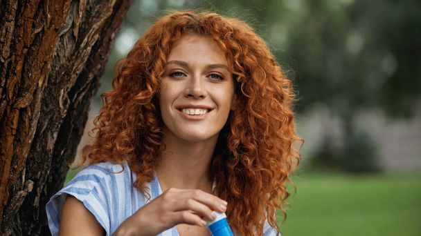 happy redhead woman holding bottle with soap bubbles in park  - Fotó, kép