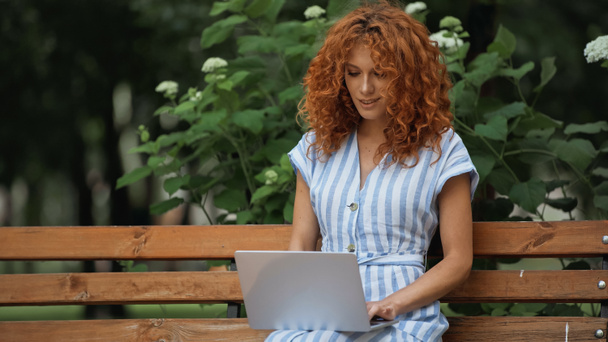 curly redhead woman smiling using laptop while sitting on bench in park  - Foto, imagen