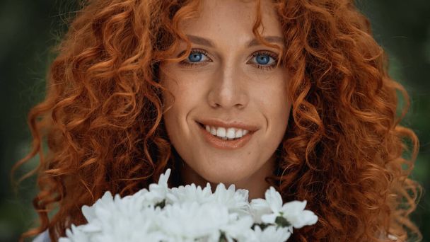 close up de encaracolado ruiva mulher com olhos azuis sorrindo perto de flores brancas  - Foto, Imagem