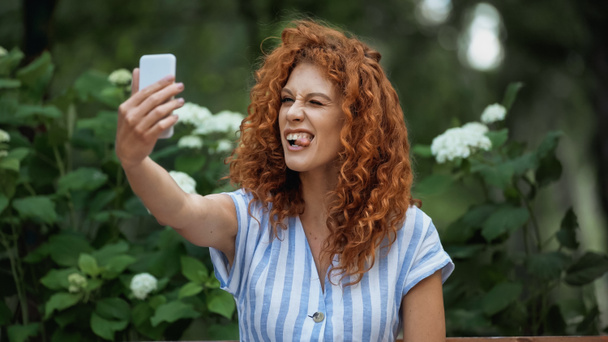 funny redhead woman sticking out tongue while taking selfie in green park  - Фото, изображение