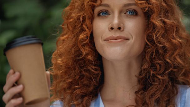 positive redhead woman with blue eyes holding paper cup outside  - 写真・画像