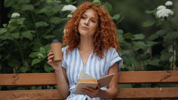 pleased redhead woman in striped dress holding book and paper cup while sitting on bench in park  - Фото, изображение