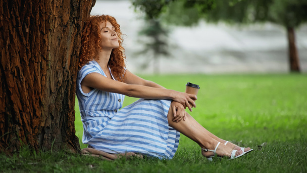 smiling redhead woman in blue dress sitting under tree trunk and holding coffee to go  - Zdjęcie, obraz