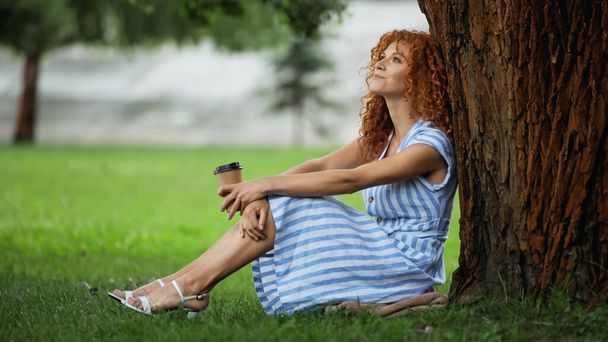 joyful redhead woman in blue dress sitting under tree trunk and holding coffee to go  - Photo, Image