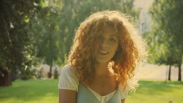 luz do sol no cabelo encaracolado vermelho da mulher sorrindo enquanto olha para a câmera no parque - Foto, Imagem