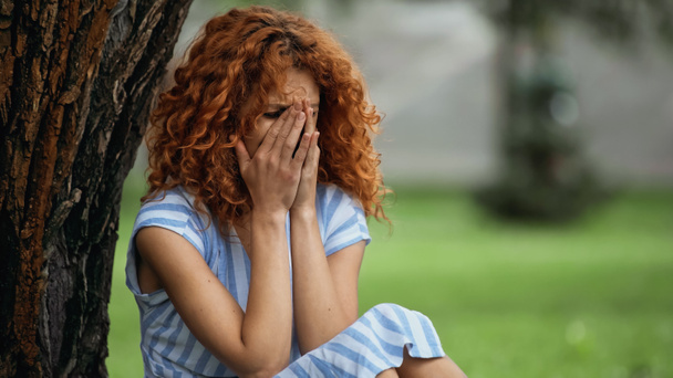 triste joven con el pelo rojo cubriendo la cara mientras llora en el parque  - Foto, Imagen