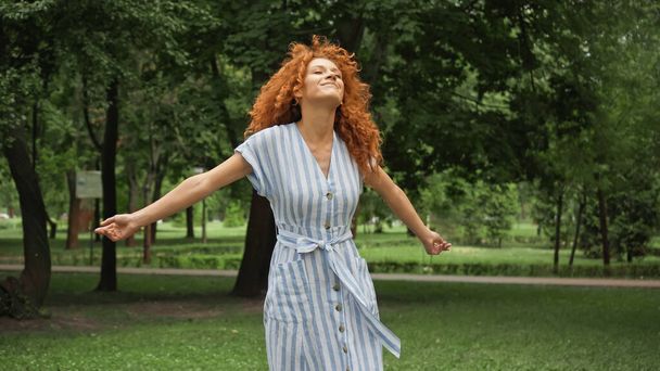 緑の公園で手を伸ばして立つ赤い髪の若い女性  - 写真・画像
