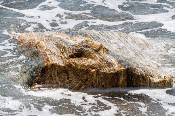 Una hermosa piedra grande y colorida está en la playa junto al mar azul con olas y espuma - Foto, Imagen