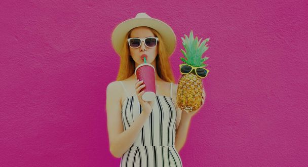 Portrait of stylish woman drinking a juice with funny pineapple wearing a summer round straw hat posing on a colorful pink background - Фото, зображення