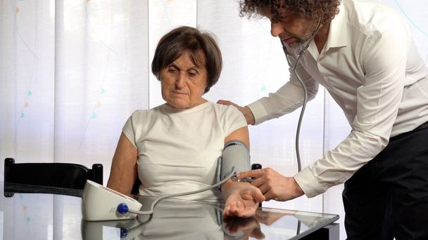 Arzt misst den Druck einer älteren Frau mit einem Tonometer - Privatarztbesuch in der Wohnung während der Covid-19 Coronavirus Lockdown Epidemie - Foto, Bild