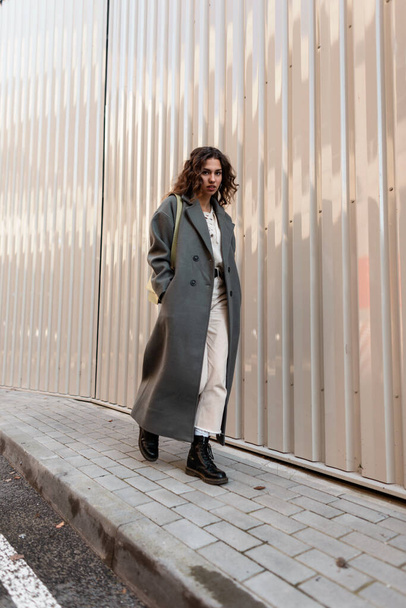 Élégante fille hipster urbaine aux cheveux bouclés dans un long manteau vert à la mode avec un sac et des bottes se promène dans la rue près d'un mur métallique - Photo, image
