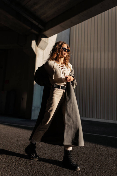 Hübsches junges, lockiges Mädchen mit Sonnenbrille in modischer Kleidung trägt einen langen Mantel, der im Sonnenlicht durch die Stadt spaziert. Femininer urbaner Stil und Schönheit - Foto, Bild