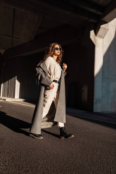 Fashionable beautiful woman with curly hair in a vintage long coat, sweater, pants and boots with sunglasses walks in the city near concrete architecture in the sunlight and shadow. - Foto, Imagen