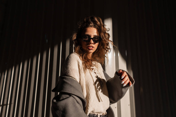 Retrato de luz solar de una hermosa joven con el pelo rizado con gafas de sol con estilo en un abrigo de moda y suéter camina por la calle cerca de una pared de metal. Estilo casual femenino y moda - Foto, imagen