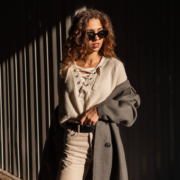 Sunny portrait of a beautiful young woman with curly hair in stylish urban clothes look with a fashionable coat and knitted blouse in sunglasses on the street - Foto, Bild