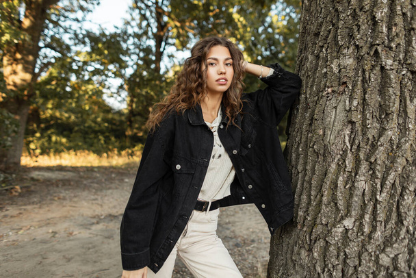 Pretty American model girl with curly hair in a denim black jacket with a sweater stands and look at camera near a tree in nature. Summer casual feminine style and beauty - Fotó, kép