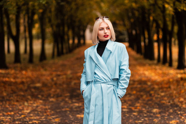 Fashionable beautiful young business woman in a stylish blue coat walks in the autumn park with yellow fall leaves - Photo, Image