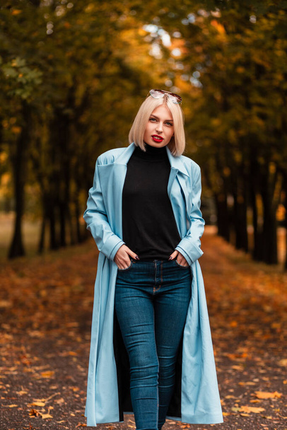 Jolie femme blonde aux lèvres rouges en manteau bleu mode, pull noir et jean se promène dans le parc d'automne. Automne style féminin et beauté - Photo, image