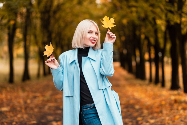 Beautiful happy young stylish girl in fashionable clothes with a blue coat, sweater and jeans holds yellow autumn leaves on the background of a fall park - 写真・画像